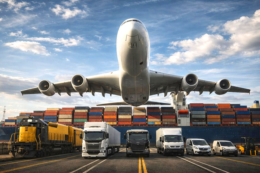 multiple transportation vehicles in front of shipping containers with airplane flying above